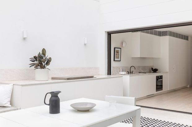 Bright, minimalist open-plan interior showing a white dining table in the foreground and a sleek white kitchen beyond. The kitchen features flat-panel cabinetry, a pale stone benchtop and splashback, integrated appliances, a black tap and sink, and light timber flooring. A large opening with dark trim connects the kitchen to the dining/living area, while sparse furnishings and white walls create an airy, calm atmosphere.
