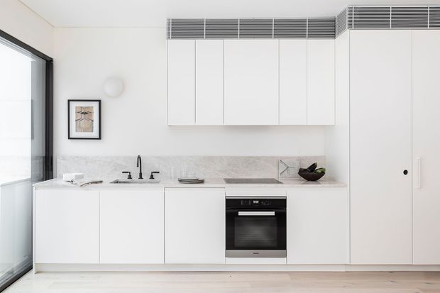 A bright, minimalist kitchen with white full-height cabinetry, a pale stone benchtop and splashback, integrated black oven and cooktop, black tapware, and a large glazed door or window at the left. Linear louvred ventilation grilles run along the upper wall and ceiling junction, while light timber flooring and restrained artwork add warmth.