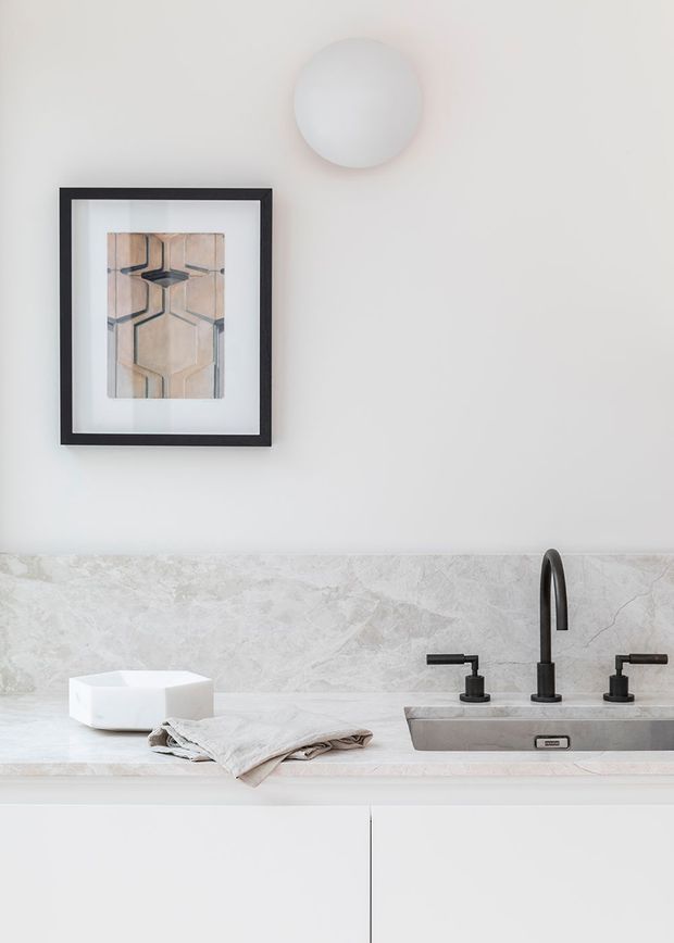 Minimal, refined bathroom vanity vignette with a pale stone countertop and integrated rectangular basin, dark metal wall-mounted-style tapware, white flat-panel cabinetry, and a light grey marble-look backsplash. A framed geometric artwork and circular wall light add restrained decorative detail.