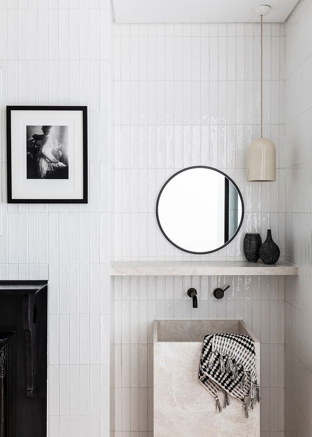 A compact, refined bathroom vanity area with glossy white vertical wall tiles, a pale stone floating shelf and basin, matte black wall-mounted tapware, and a round black-framed mirror. A sculptural pendant light, monochrome artwork, and black decorative vessels add contrast. The edge of a dark fireplace or architectural feature is visible at left.