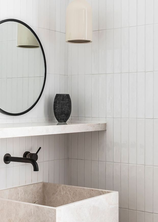 Close-up of a refined bathroom vanity featuring vertically laid white wall tiles, a pale stone vessel basin, dark wall-mounted tapware, a floating stone shelf, a round black-framed mirror, and minimalist pendant lighting. A dark woven decorative vessel adds contrast.