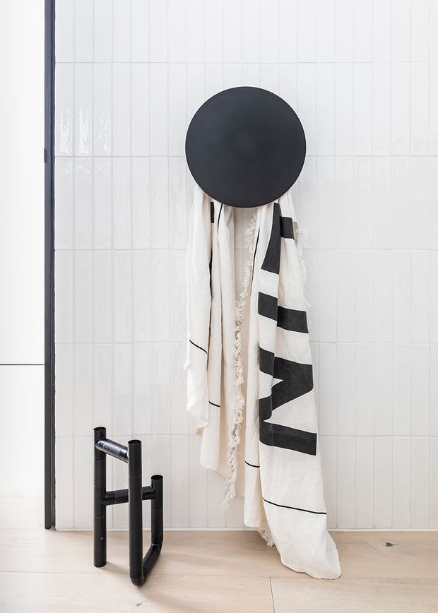 Minimalist bathroom detail featuring glossy white vertical wall tiles, a large round matte-black wall fixture or mirror, monochrome hanging textiles, and a sculptural black freestanding tap fitting beside a pale timber-toned floor.