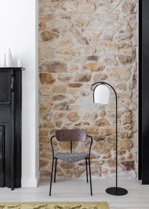 Interior vignette featuring a full-height exposed natural stone masonry wall in warm beige and brown tones, framed by white plaster walls and dark painted timber elements. A mid-century-inspired chair with a dark wood back and patterned upholstered seat sits beneath a slim black floor lamp with a white ribbed shade. Pale timber flooring and a partial area rug complete the refined, textural setting.
