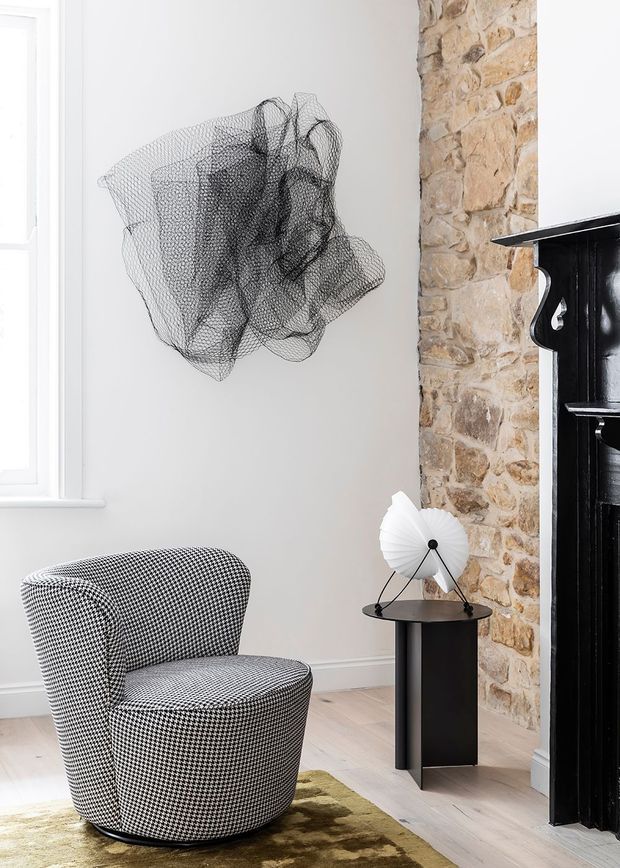 A refined sitting area featuring a sculptural black-and-white patterned lounge chair on a muted rug, beside a slim black pedestal table with a pleated white lamp. A rough exposed stone chimney breast and glossy black traditional fireplace surround contrast with the smooth pale walls, timber floor, and abstract wire wall sculpture. Natural light enters through a tall window.