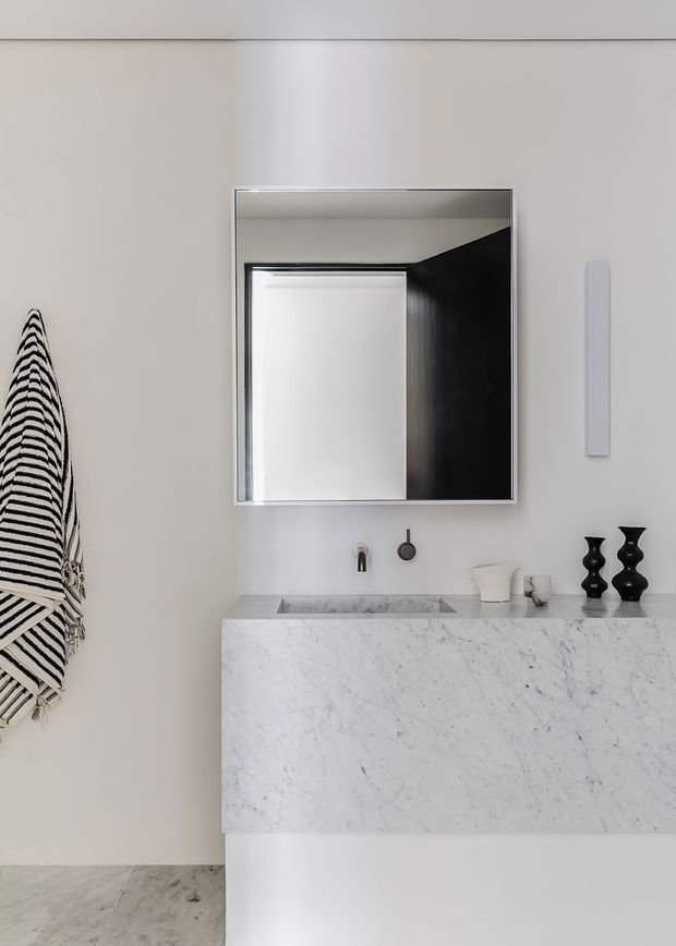 A minimalist bathroom vanity with a thick white marble basin and front panel, wall-mounted black tapware, a large rectangular mirror, and black decorative vessels. A striped black-and-white textile hangs on the left wall, with pale neutral walls and a light stone floor.