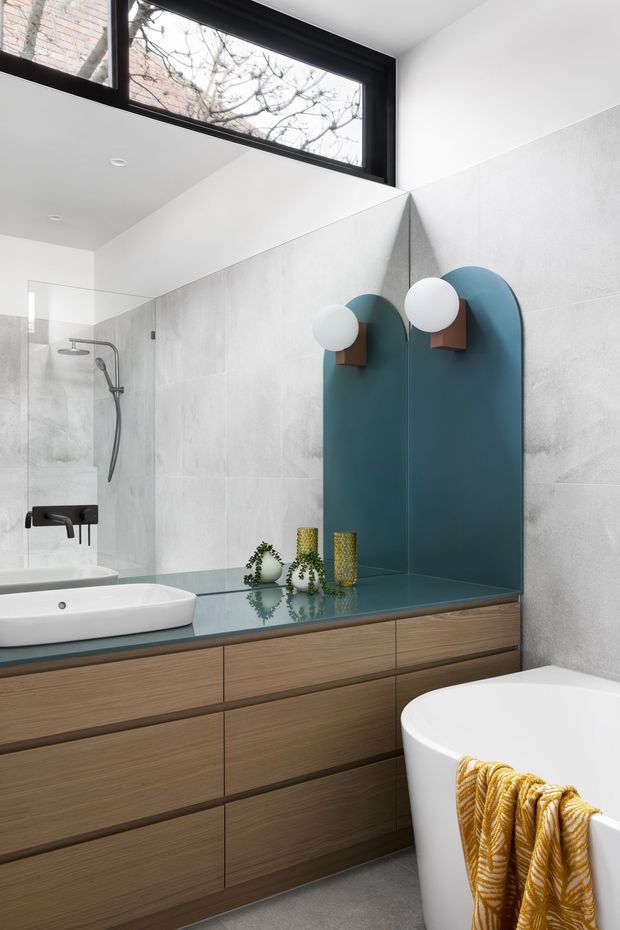 Contemporary bathroom with a long floating timber vanity, white vessel basins, dark teal glass benchtop and matching arched mirror panels. The walls are clad in pale grey textured tiles, with a frameless glass shower enclosure, brushed metal shower fittings, sculptural globe wall lights, and a freestanding white bathtub. High-level black-framed glazing brings in natural light, while a mustard patterned towel adds colour.