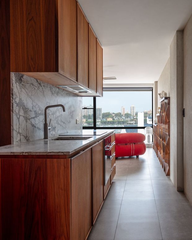 A narrow, contemporary galley kitchen with rich dark timber cabinetry, a dramatic veined stone backsplash and benchtop, and a sculptural dark metal faucet. The kitchen opens toward a bright living area with floor-to-ceiling glazing and a city or waterfront skyline beyond. A vivid red rounded lounge chair and timber shelving add contrast, while large pale floor tiles and smooth plaster walls create a refined, warm-modern interior.