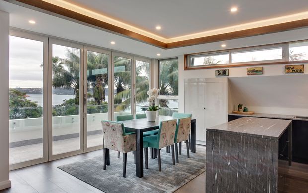 Bright contemporary open-plan kitchen and dining area with floor-to-ceiling glazing and multiple glass doors opening to a balcony. The room features a dark stone kitchen island, white cabinetry, a teal dining table, upholstered dining chairs, recessed lighting, and a timber ceiling perimeter detail. Tropical palms and a waterfront view are visible outside.