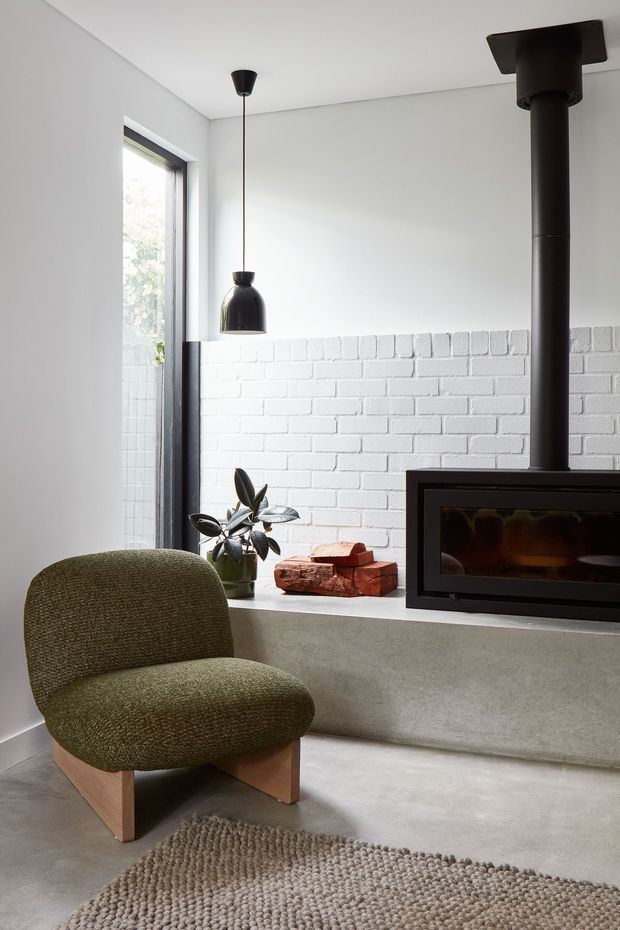 A minimalist living room featuring a freestanding black wood-burning fireplace with a tall flue, set into a low concrete hearth against a white-painted brick wall. The room has polished grey concrete flooring, a narrow black-framed window, a sculptural pendant light, and a low olive-green upholstered armchair on timber legs. Firewood and a potted plant sit on the hearth, with a textured neutral rug in the foreground.