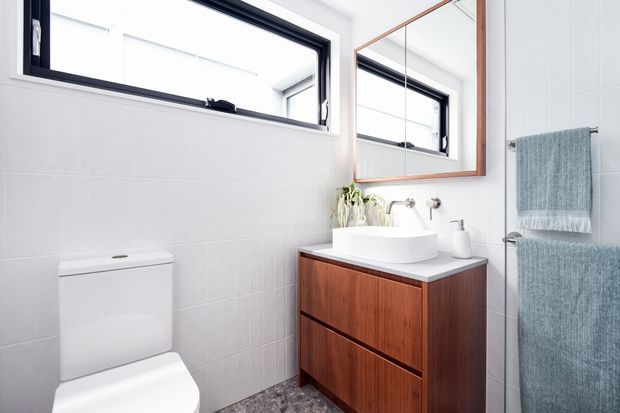 Bright, compact contemporary bathroom featuring white tiled walls, a wall-mounted black-framed window, timber vanity with a white vessel basin, large mirrored cabinet with timber trim, chrome fixtures, and a glass shower enclosure with pale blue towels.