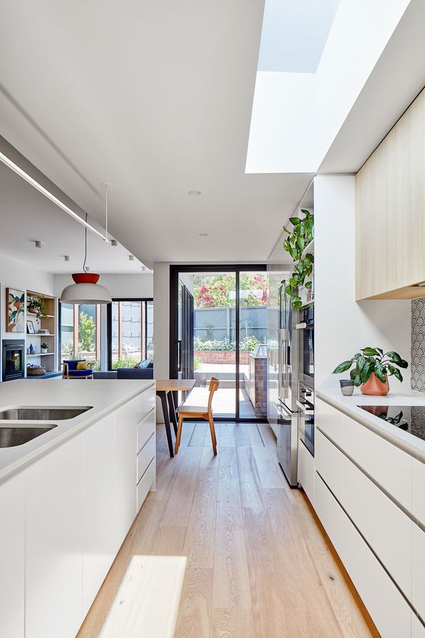 Bright contemporary open-plan kitchen with white handleless cabinetry, a large central island with integrated sink, pale timber flooring, dark-framed glazing, and a dining nook leading to an outdoor courtyard. A long skylight brings strong natural light into the space. The right-hand cabinetry includes a patterned tile splashback and open shelving with plants.