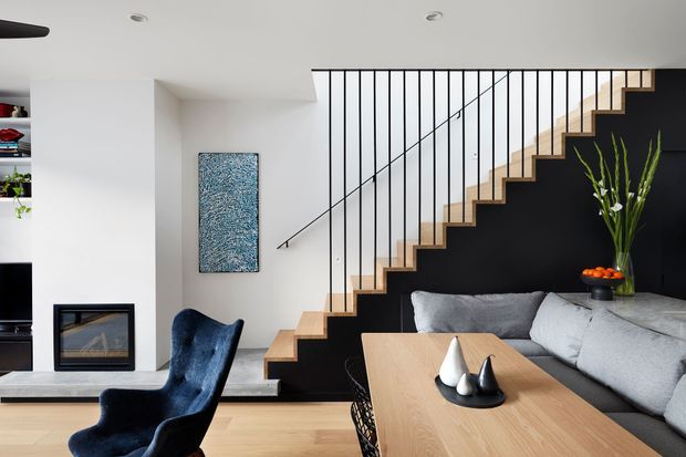 Contemporary open-plan living and dining area with a dramatic floating timber staircase set against a matte black feature wall. The room includes a large light-grey sectional sofa, timber dining table, dark upholstered dining chair, integrated fireplace, shelving, abstract artwork, and a tall indoor plant. White walls and ceiling, pale timber flooring, and black vertical stair balusters create a clean, refined contrast.