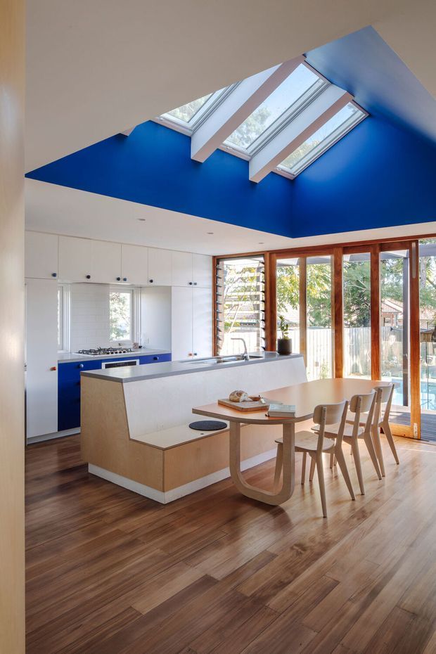Bright open-plan kitchen and dining area with a central island, white built-in cabinetry, timber flooring, and a light timber dining table. A vivid cobalt-blue ceiling recess surrounds three skylights, while large timber-framed glazed doors and windows provide abundant natural light and views to the outdoors.