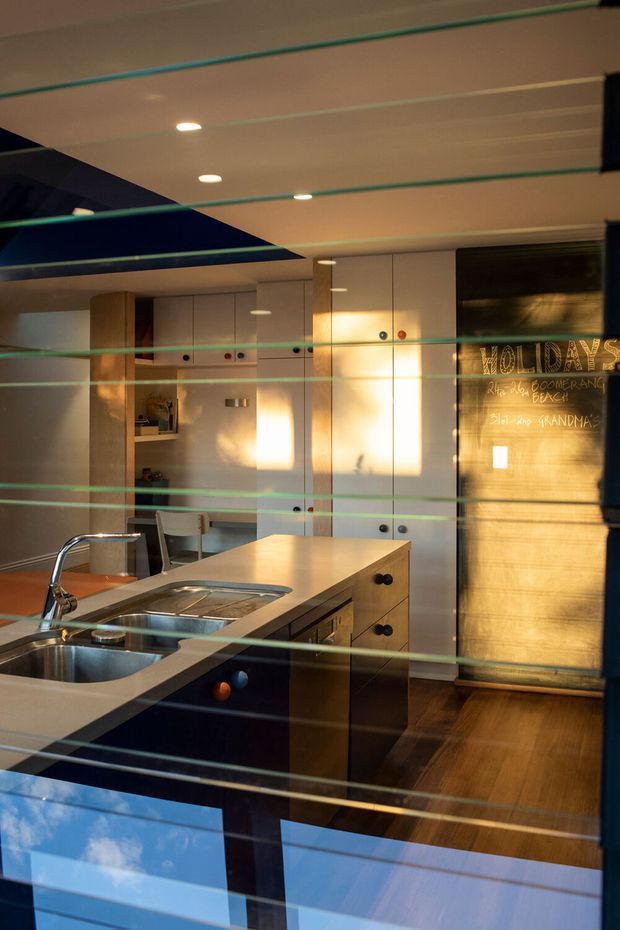 A contemporary kitchen viewed through reflective glass, with a central island featuring an integrated double sink and chrome tapware, pale cabinetry, dark lower joinery, timber flooring, and warm sunlight. A chalkboard-style wall panel with handwritten notes forms a feature surface on the right. Reflections and horizontal lines from the glazing overlay the scene.
