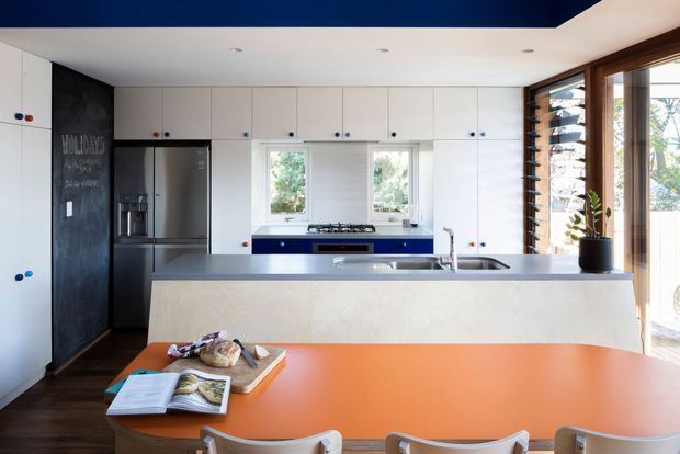 A bright contemporary kitchen viewed from the dining area, featuring a large orange dining table in the foreground, white flat-panel cabinetry, a dark integrated refrigerator wall, blue accents beneath the cooktop, pale stone-look island benchtop, stainless-steel sink and tap, and timber-framed louvre windows along the right side. Natural light enters through the windows and glazed doors.