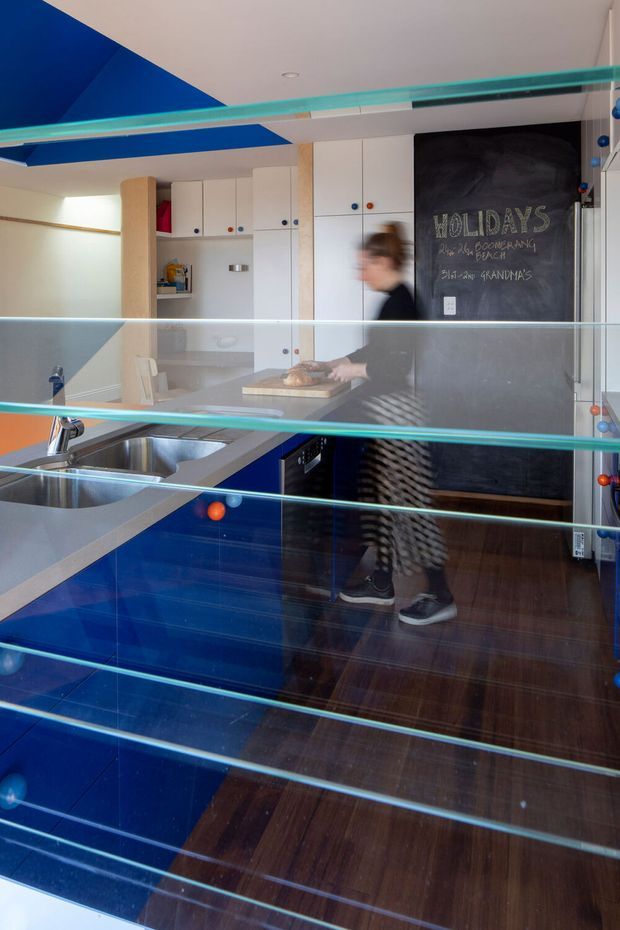 A compact, highly graphic modern kitchen viewed through a glass balustrade or partition. The space features vivid cobalt-blue cabinetry and ceiling accents, white built-in cupboards, a dark chalkboard wall, stainless-steel double sink, pale benchtop, and dark timber flooring. A person is moving through the kitchen, creating motion blur. Horizontal glass rails dominate the foreground.