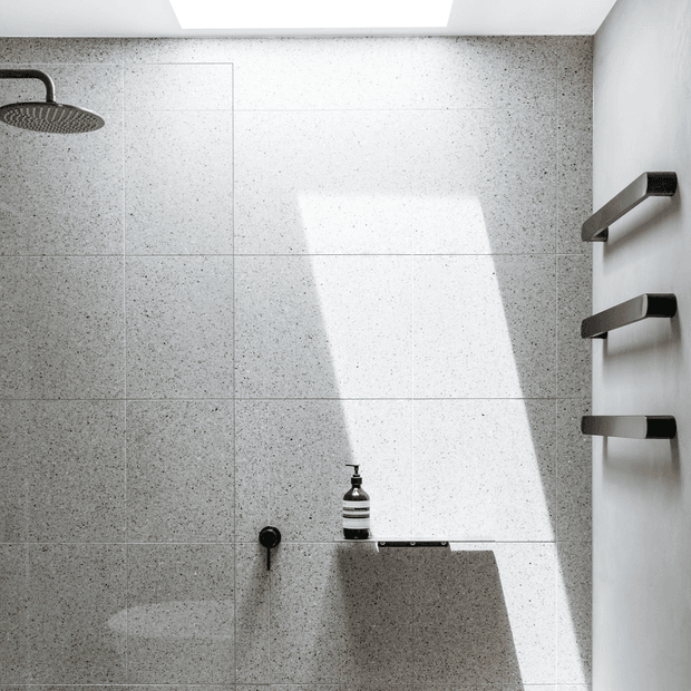 Minimalist contemporary walk-in shower with large speckled grey stone-look wall tiles, matte dark shower fittings, three wall-mounted towel rails, and a recessed ceiling skylight casting strong natural light across the shower wall.