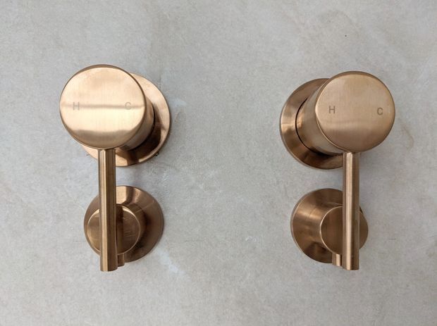 Close-up of two wall-mounted cylindrical hot-and-cold water controls in a bathroom, finished in warm brushed bronze or brass-toned metal against a light grey textured wall. The minimalist round escutcheons and lever handles form a symmetrical, refined fixture composition.