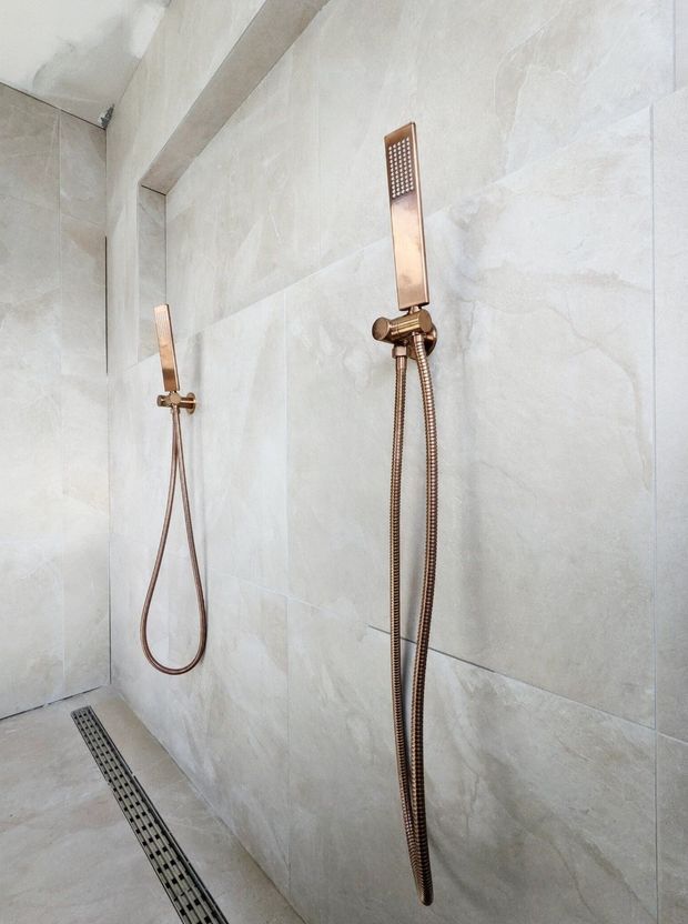 A minimalist walk-in shower with large pale grey stone-look wall and floor tiles, two wall-mounted handheld shower sets, warm bronze fixtures, and a long linear floor drain. The space is clean, contemporary, and softly lit.
