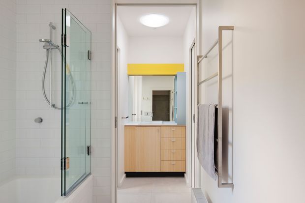Bright contemporary bathroom viewed toward a vanity area. The room features a frameless glass walk-in shower with white wall tiles, a floating light-wood vanity with drawers, a large mirror, white surfaces, and a wall-mounted towel rail with folded towels. A vivid yellow horizontal accent panel adds colour near the vanity.