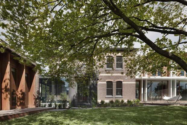 A landscaped courtyard view of a refined multi-building residential property. A warm timber-clad structure with large vertical doors sits to the left, connected visually to a glazed pavilion and a pale brick multi-storey building with dark-framed windows and a covered outdoor terrace. Mature tree canopy and lawn frame the composition.