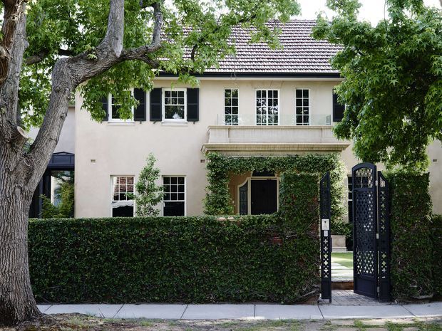 Front exterior view of a two-storey traditional residence, partially screened by mature trees and a neatly clipped hedge. The pale rendered facade has a tiled pitched roof, multi-pane sash-style windows with dark shutters, a projecting decorative entry canopy, climbing greenery, and an ornate black pedestrian gate leading to the front door.