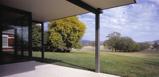 View from a covered outdoor terrace or patio toward a broad landscaped lawn and open rural landscape. The space has a flat roof canopy supported by dark square posts, pale concrete paving, and large dark-framed glazed doors at the left. Mature trees, distant hills, and a low red-brick building are visible beyond the property.