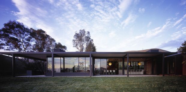 A low, single-storey modern residence viewed from the lawn, with extensive floor-to-ceiling glazing, dark slender framing, broad flat roof planes, and a partially enclosed covered outdoor area. Interior dining and living spaces are visible through the glass, while mature trees and open landscape surround the home.