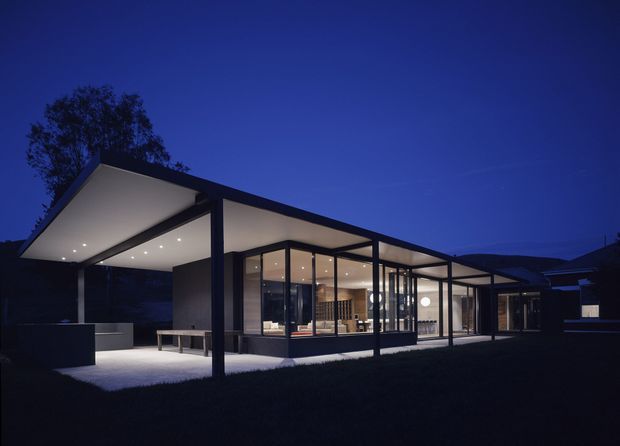 A low, single-storey modern residence photographed at dusk, with broad flat roof planes, slender dark structural posts, extensive floor-to-ceiling glazing, and a covered outdoor terrace or carport-like canopy. Warm interior lighting reveals an open-plan living area and fixed kitchen joinery behind the glass. The building is set against a dark landscaped site with silhouetted trees and distant hills.
