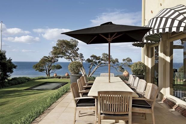 A spacious seaside terrace overlooking the ocean, furnished with a long outdoor dining table, wooden slatted dining chairs, and a large dark cantilever-style umbrella. The terrace adjoins a light-coloured building with striped fabric awnings and glazed doors. A manicured lawn, landscaped planting, and a narrow dark reflecting pool or water feature extend toward the coastal view.