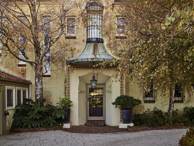 Exterior view of an ornate historic-style stucco residence, centered on a recessed entry with a decorative metal security door, lantern, patterned tile trim, and a distinctive curved metal canopy topped by a tall enclosed cupola. Mature trees and clipped shrubs frame the facade, with a gravel forecourt and brick edging in front.