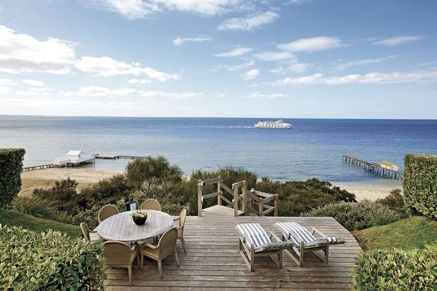 A coastal outdoor deck overlooking a calm ocean, with a round outdoor dining table and chairs, two striped sun loungers, timber steps descending through landscaped coastal vegetation, sandy beaches, and small jetties on either side. The scene is bright and relaxed with a wide ocean horizon and partly cloudy sky.