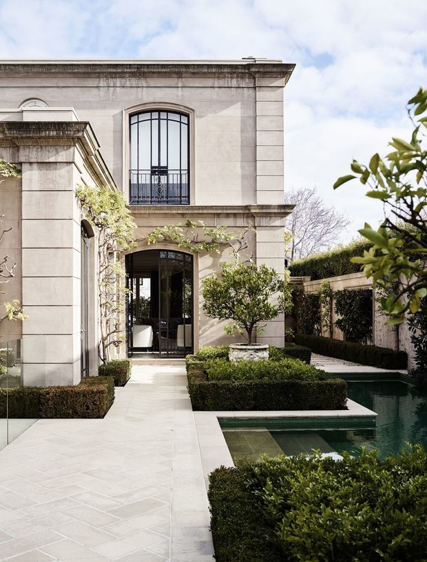 A formal courtyard garden beside an elegant two-storey rendered masonry building. The facade features large arched steel-framed windows and doors, classical horizontal banding, cornices and pilasters. Carefully clipped hedges, espaliered greenery and a rectangular reflecting pool line a pale geometric stone-paved walkway.