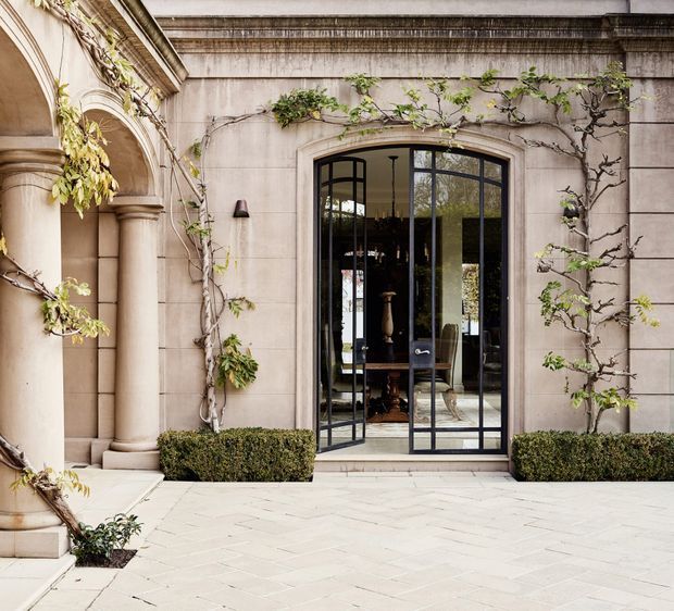 A refined stone courtyard facade with classical architectural detailing, including an arched colonnade, pilasters, moulded cornice, and a tall black metal-framed glazed doorway. Climbing vines cover parts of the pale stone walls, with clipped hedges and pale herringbone paving at the entrance. A dark, traditionally styled interior is visible through the open doors.