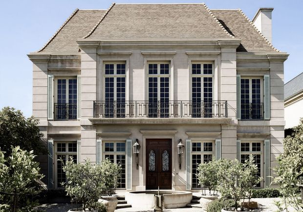 Front exterior view of a symmetrical, stately two-storey residence with a pale rendered masonry facade, tall multi-pane French-style windows, ornate dark metal balcony railings, shuttered side windows, and a steep hipped slate-like roof. A central dark timber entrance is approached by steps and framed by formal landscaping and a shallow stone water feature.