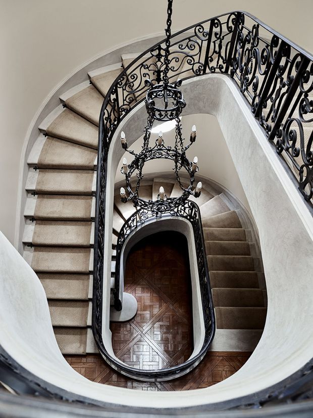 A dramatic view down an ornate curved stairwell with multiple winding flights, cream carpeted steps, white plaster walls, elaborate black wrought-iron balustrades, and matching multi-tier chandeliers. A patterned dark timber parquet floor is visible at the lower level, emphasizing the vertical void and elegant historic detailing.