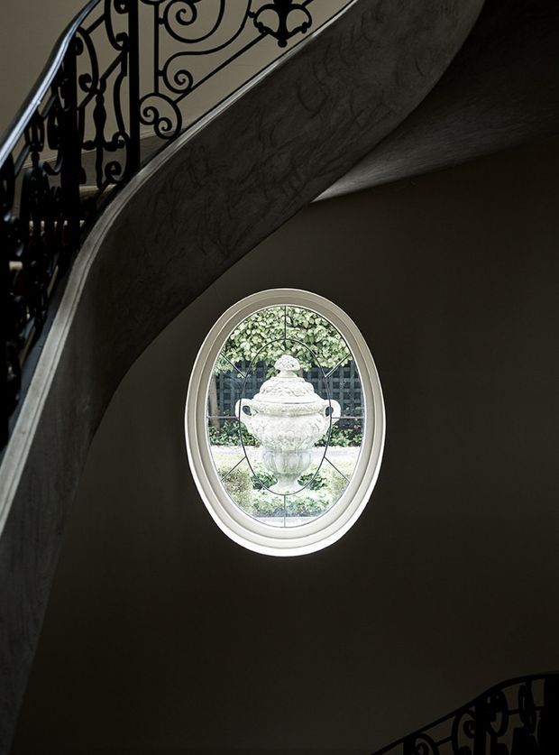 A dramatic interior view of a curved stairway with ornate black wrought-iron balustrades and a sculptural sweeping underside. A small oval decorative window with layered white trim and patterned glazing overlooks a garden and urn outside. The dark walls and sculpted stair form create a grand, traditional atmosphere.