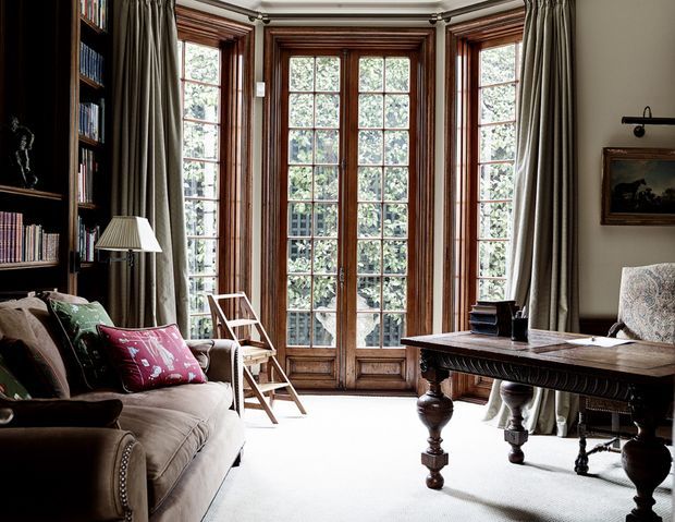 Elegant traditional library or study with a large projecting bay of tall multi-pane timber-framed glazed doors and windows, framed by full-length taupe curtains. A dark built-in bookcase lines the left wall beside a plush sofa with patterned cushions, while a substantial dark timber trestle-style desk or table occupies the right foreground. The room has ornate ceiling cornices, a pale carpet, classic furnishings, and abundant natural light from the glazing.