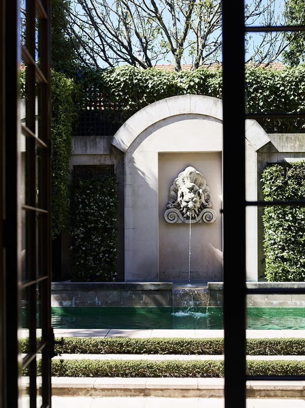 View from indoors through dark-framed glazing onto a formal courtyard garden. A rectangular green-tiled reflecting pool features a pale stone arched niche with an ornate sculptural fountain, surrounded by clipped hedges, masonry walls, and dense greenery. The scene has a classical, symmetrical, refined character with strong sunlight and shadows.