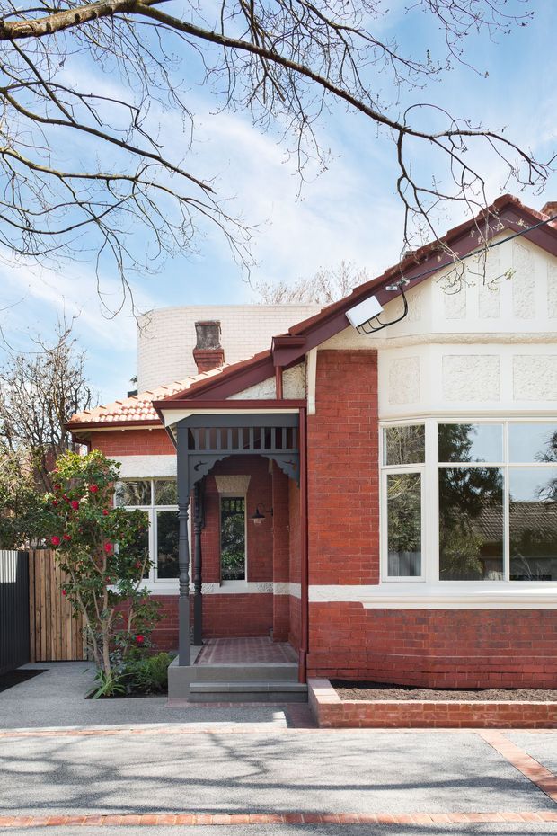 Street-facing view of a restored red-brick period house with a projecting gabled roof, cream rendered upper facade, white-framed bay window, tiled porch roof, decorative timber porch posts and brick-paved entry. Mature deciduous branches and a small planted garden frame the residential facade.