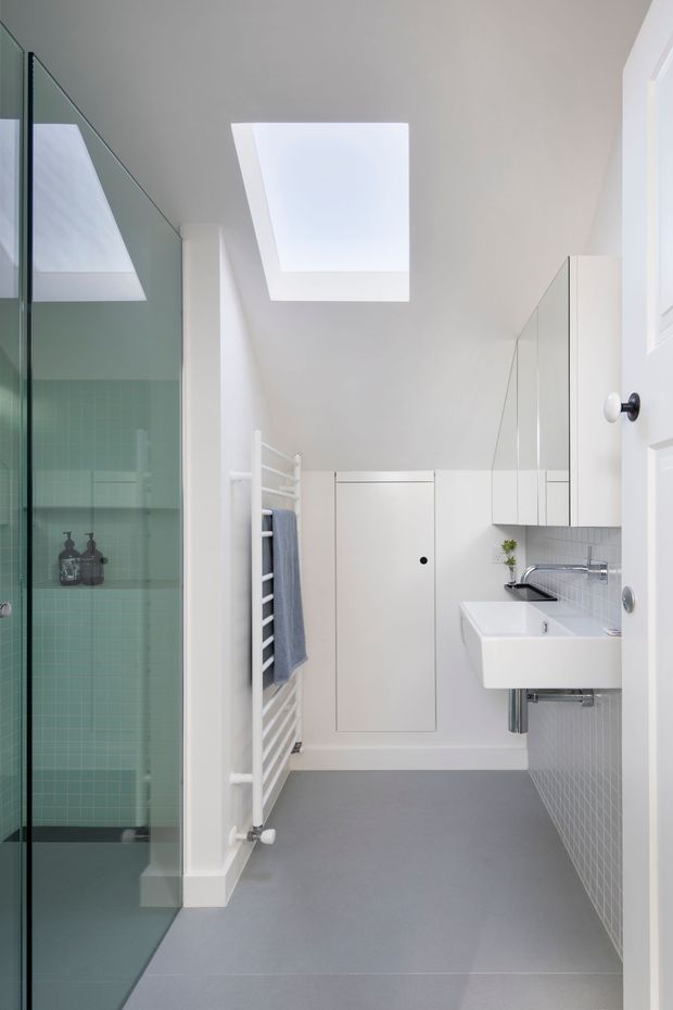 Bright, narrow contemporary bathroom with a frameless glass shower enclosure on the left, pale green square wall tiles inside the shower, a white wall-mounted basin and mirrored cabinetry on the right, a white heated towel rail, grey floor tiles, and a large skylight. The space has a clean, minimalist fit-out with abundant natural light.