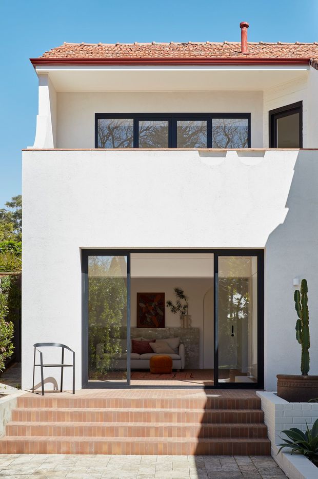 Sunlit rear exterior of a two-storey rendered house with a terracotta tiled roof, dark-framed glazed doors and windows, and a deep upper balcony or terrace. Broad terracotta tiled steps lead to a large sliding glass opening revealing a living room inside. Potted cactus and planted borders frame the patio, while the white stucco facade and strong shadows create a clean Mediterranean feel.