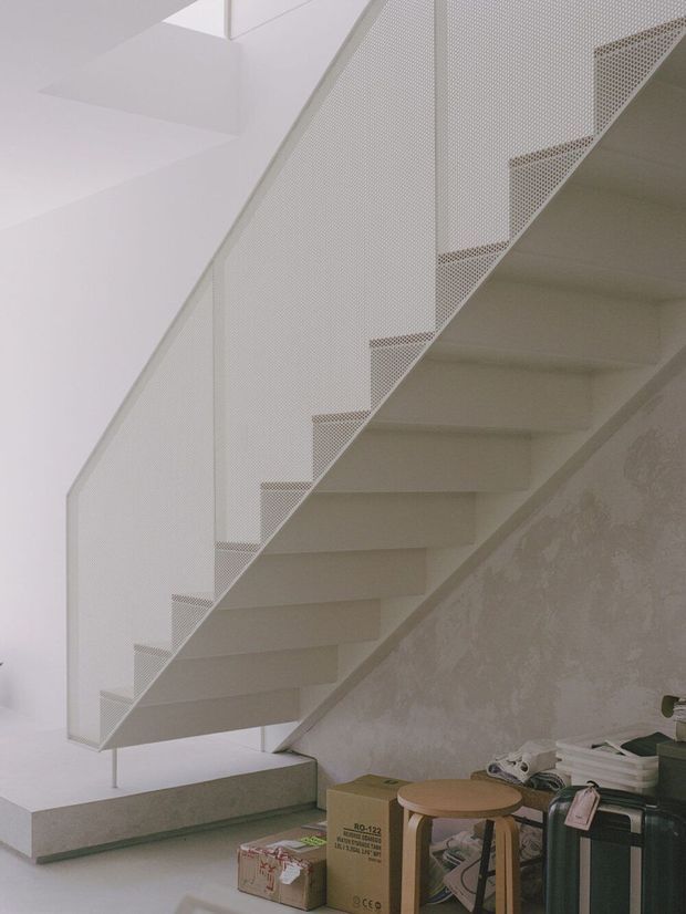 A minimalist interior view of a modern floating staircase with crisp white steps and a perforated white metal balustrade. The staircase rises diagonally through a pale, refined space with a textured light wall beneath it. Moving boxes, a stool, and luggage are visible at the base, suggesting an unfinished or recently occupied interior.