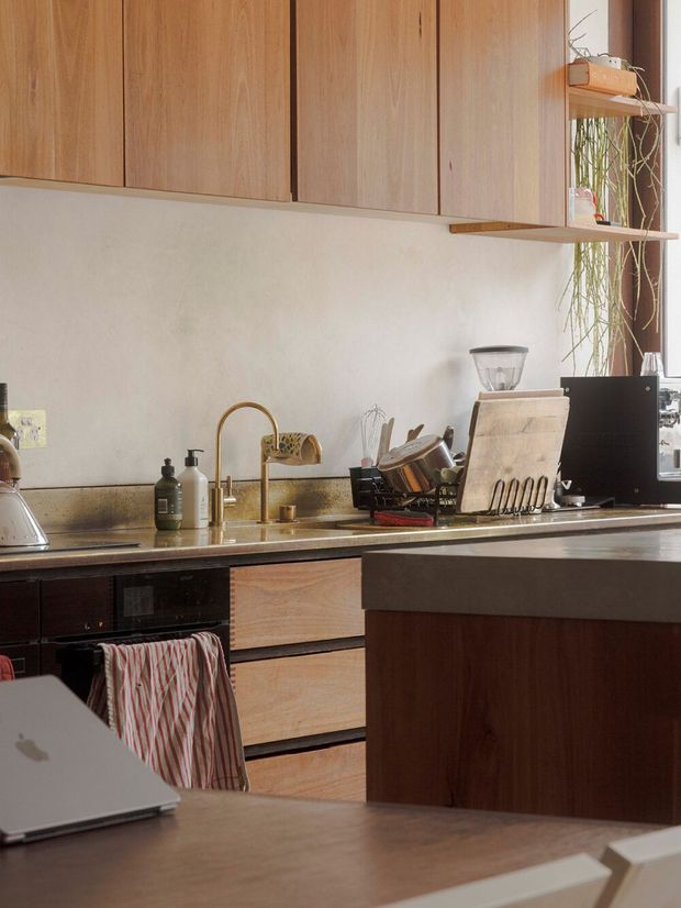 Warm, lived-in contemporary kitchen with extensive light timber overhead cabinetry, dark lower cabinetry, a pale stone or composite benchtop, brass-toned faucet and sink, open shelving, integrated appliances, and a central island. A dining table and laptop are visible in the foreground, while plants and kitchen accessories add an organic domestic feel.