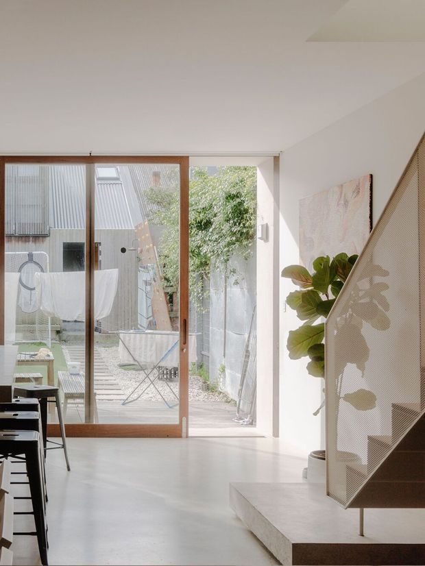 Bright, minimalist residential interior viewed from an open-plan living or dining area. A large timber-framed glazed sliding door opens to a compact courtyard, while a sculptural stair with a perforated or mesh balustrade occupies the right side. The space has white walls and ceiling, a pale smooth floor, sparse dark dining furniture, indoor planting, and abundant natural light. The courtyard and adjacent exterior are visible through the glazing.