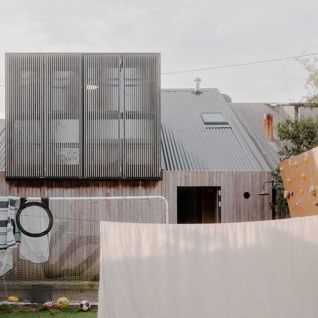 Exterior view of a compact contemporary dwelling with a corrugated metal pitched roof, timber-clad lower facade, and a prominent vertical slatted screen enclosing an upper-level section. A roof skylight and chimney are visible, with a modest outdoor recreation area and laundry in the foreground.