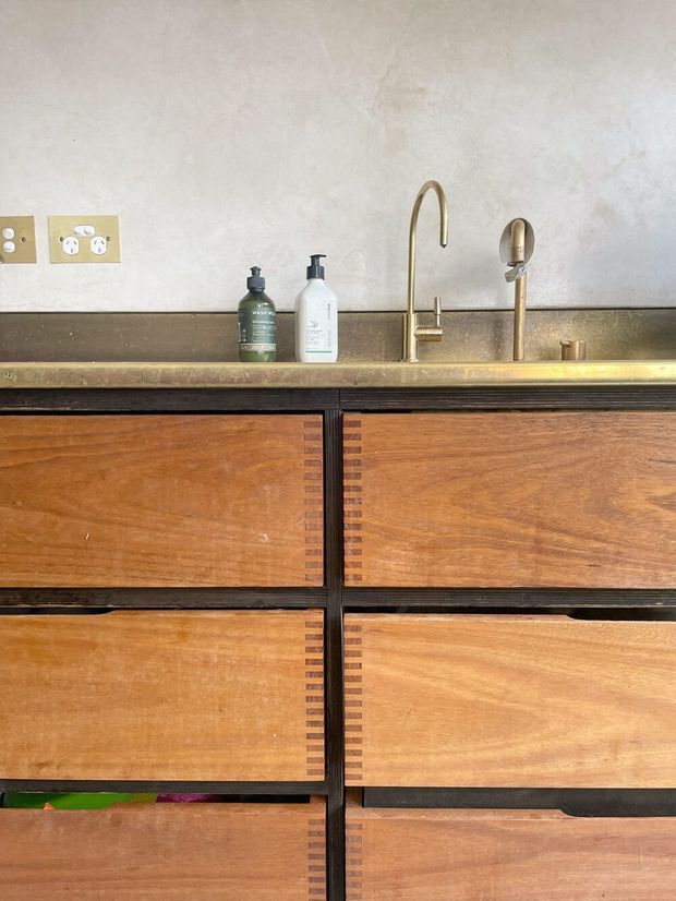 Close-up of a refined bathroom vanity with warm timber drawer fronts framed by dark detailing and decorative joinery, topped with a brass-toned benchtop or integrated basin surround. A tall brass faucet and matching hand-shower fixture sit against a textured pale plaster wall, with brass electrical plates and minimal toiletries visible.