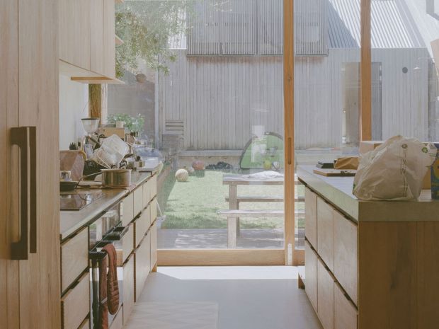 A bright, narrow galley kitchen viewed toward a full-height glazed door or window opening onto a small courtyard. Pale timber cabinetry runs along both sides, with dark stone or composite benchtops, integrated appliances, open shelving, and warm timber framing. The space has abundant natural light and a calm connection to the outdoor area.