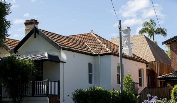Exterior view of a compact residential dwelling in a leafy suburban setting. The main house has white rendered walls, a traditional terracotta tiled gable roof, brick chimney, dark contrasting roof trims, and a small covered front porch with a striped awning and metal balustrade. A taller adjoining or neighbouring structure with a steep metal roof is visible behind it.