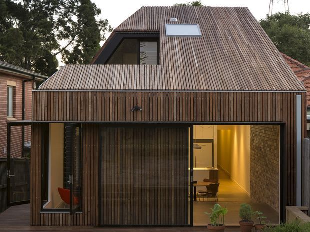 A compact contemporary residential extension viewed from the exterior, with a steeply pitched roof and facade clad entirely in narrow vertical timber battens. Large glazed openings reveal a warmly lit interior with a small dining or workspace area, while a roof skylight and upper-level window punctuate the timber roof plane. The building sits closely among neighbouring homes and mature trees.